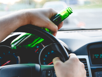 close up image of hands holding a steering wheel of a car and holding a bottle of beer