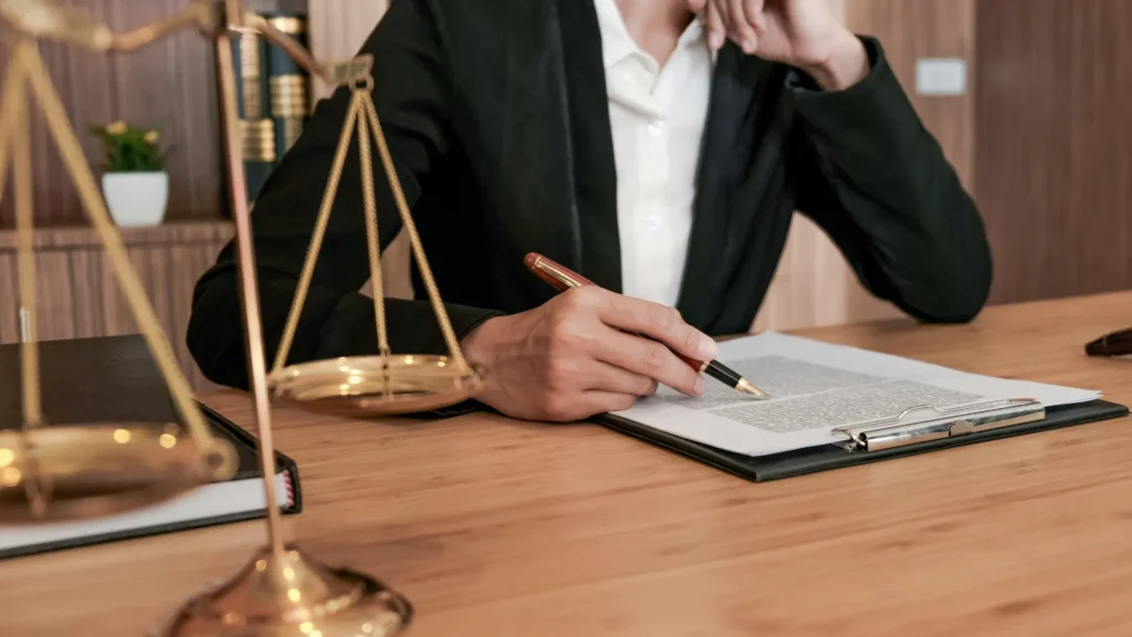 A person in a suit writing on a legal document with a pen, with a golden balance scale in the foreground, symbolizing justice and legal work.