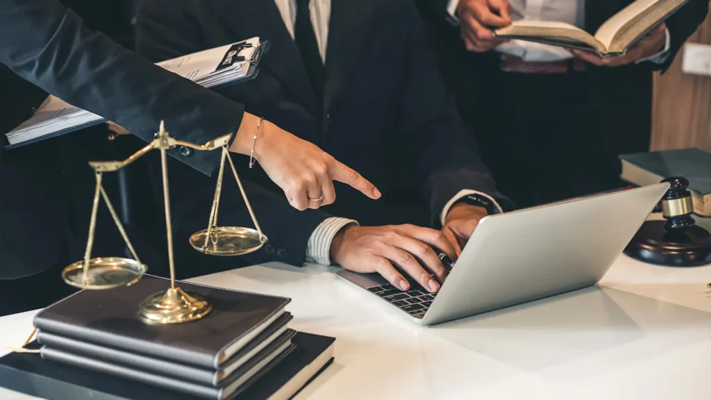 A group of legal professionals in suits collaborating at a desk with a laptop, legal books, and a balance scale, symbolizing justice and legal work