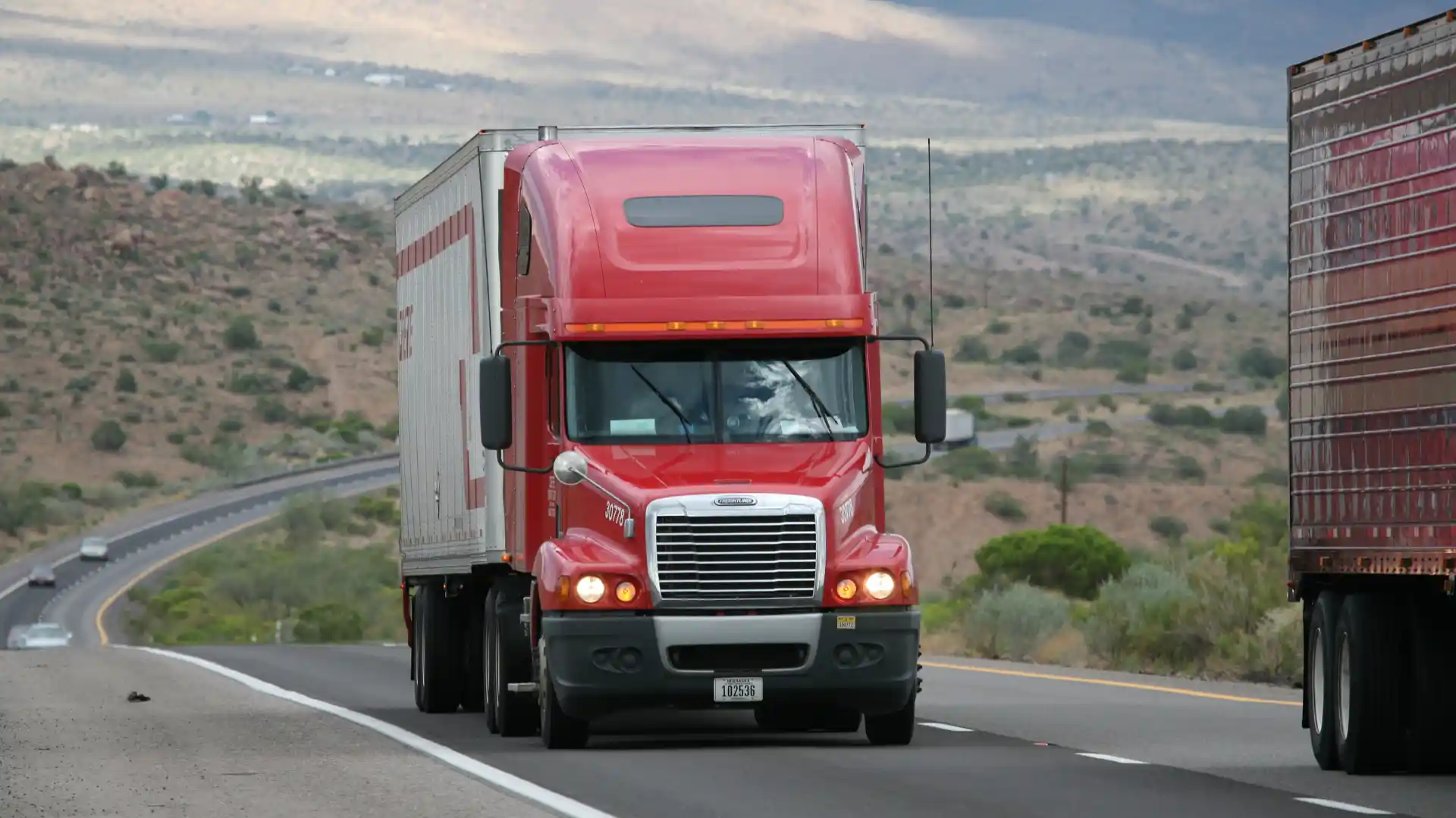 Red semi-truck driving on a highway in a rural area.