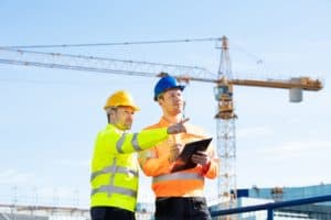 Two construction workers in safety gear discussing plans at a building site with a crane in the background.